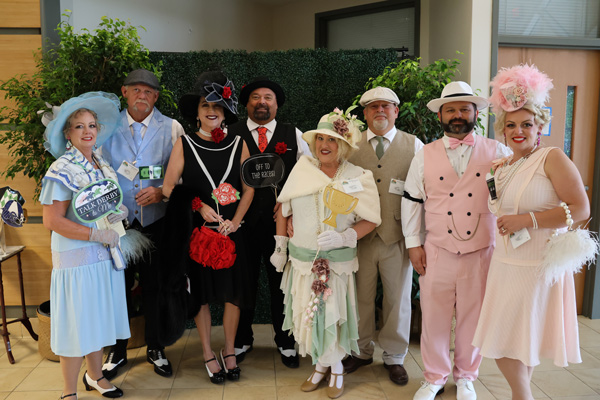8 couples smiling, dressed up in 1920's dress with hats and ties